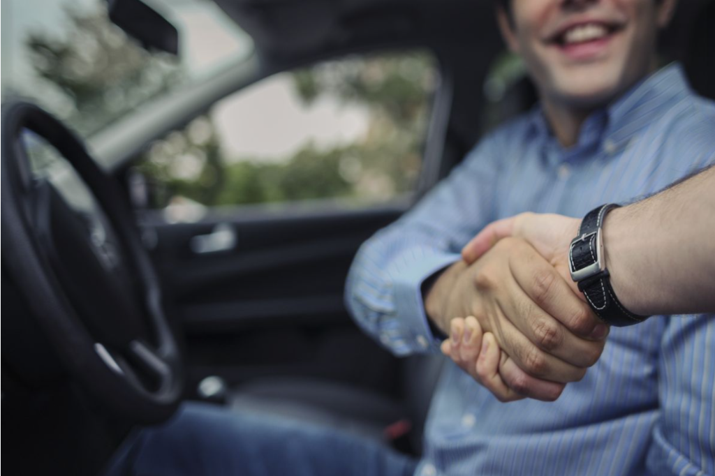A man sitting in the drivers seat of a car and shaking a car salesman hand.