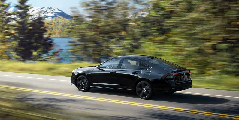 A black 2025 Honda Accord driving down a road with a lake in the backround.