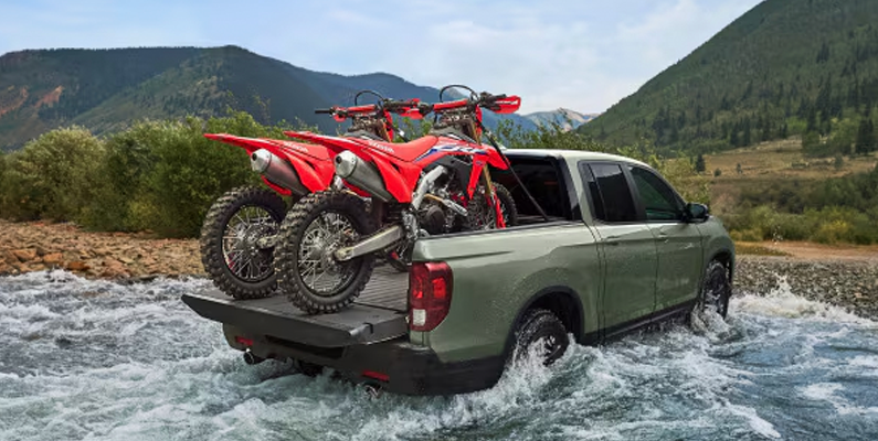 A photo of a green 2026 Honda Ridgeline driving through a stream of water with dirtbikes in the bed of the truck.