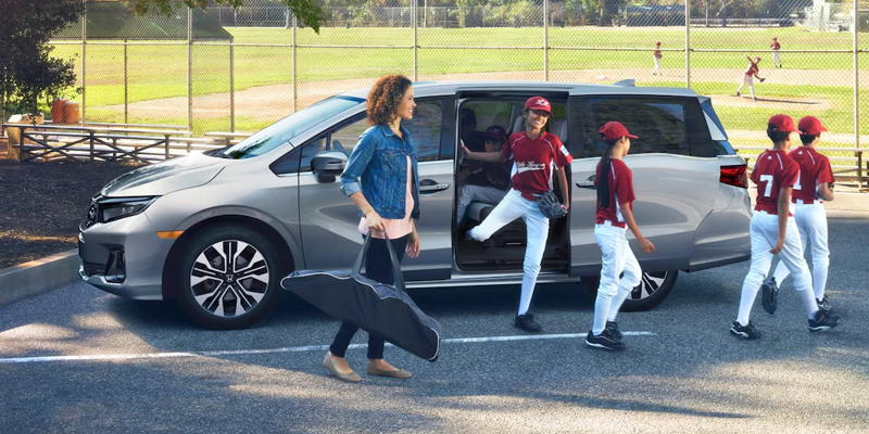 A woman walking beside a minivan as baseball players exit the vehicle, heading towards a field.